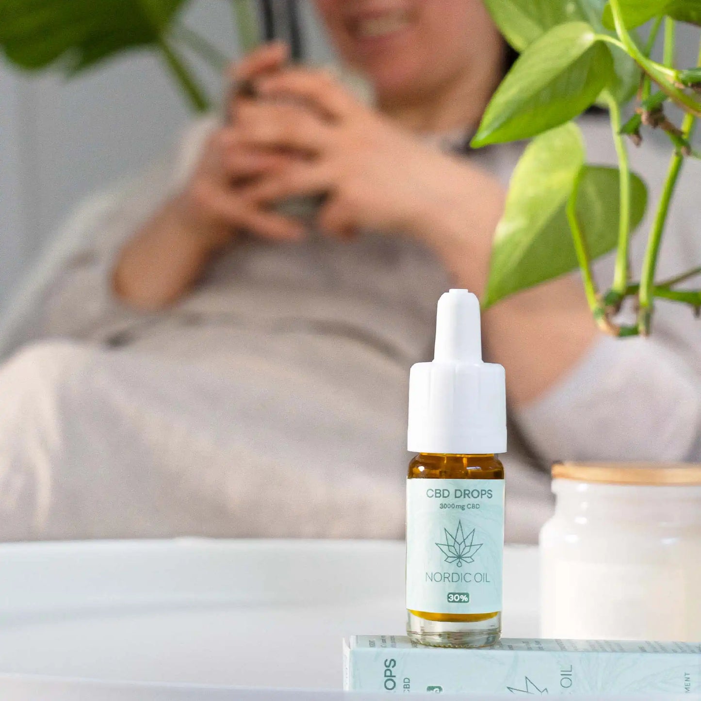 the oil is standing on a white table and in the background is a woman sitting on a sofa